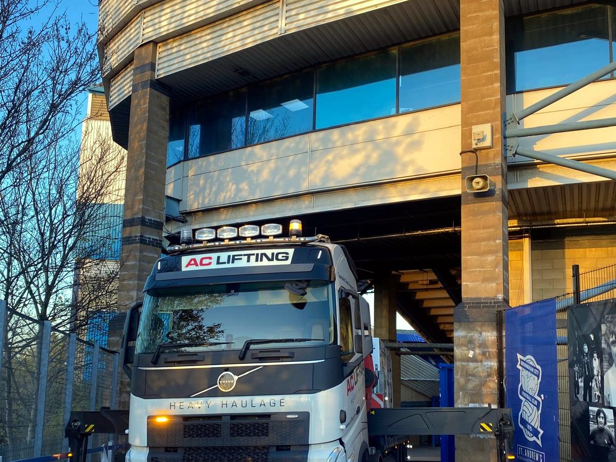 Branded AC Lifting truck driving beneath a tall industrial building.